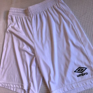 Vita träningsshorts från Umbro - Säljer ett par vita Umbro shorts i storlek M, det finns en liten fläck som syns på bild 5 som försvinner vid tvätt. perfekta för sport eller chill. De har elastisk midja och är gjorda i 100% polyester för skön och lätt känsla. Klassisk design med Umbro-logga på benet. Passar dig som gillar enkel och sportig stil.