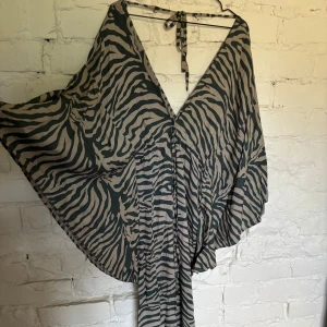 Zebramönstrad långklänning med v-ringning - Snygg långklänning med zebramönster i svart och beige. Klänningen har en djup v-ringning och vida, fladdriga ärmar som ger en cool och avslappnad vibe. Knytband i nacken för extra detalj. Perfekt för dig som gillar att sticka ut med mönster och flowy siluett.