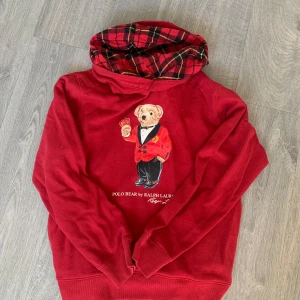 Polo bear hoodie - Säljer en röd hoodie från Ralph Lauren med Polo Bear-motiv på bröstet. Insidan av huvan har ett snyggt röd- och svart-rutigt mönster. Tröjan har långa ärmar och är gjord i mjuk bomull, perfekt för dig som gillar klassisk streetstyle med en twist. Storleken är S. Priset är inte hugget i sten.
