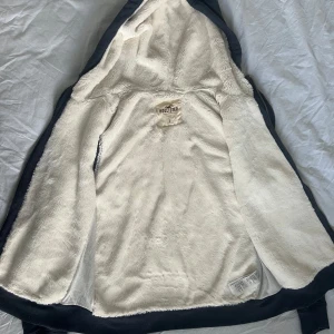 Mörkblå teddyjacka från Hollister - Mysig mörkblå faux fur teddyjacka från Hollister med fluffigt vitt foder och huva. Jackan har dragkedja framtill, snörning vid huvan och två fickor. Stort Hollister-tryck på bröstet ger en cool vibe. Perfekt för kyliga dagar när du vill vara både bekväm och snygg.
