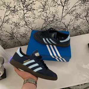 Adidas Handball Spezial mörkblå sneakers - Adidas Handball Spezial sneakers i mörkblå mocka med ljusblå ränder och detaljer. Klassisk siluett med gummisula och snörning. Ikonisk Spezial-logga i guld på sidan och Adidas-märke på plösen. Perfekt för dig som gillar retrostil och streetwear. Köpta från Zalando Max använda 15 gånger.