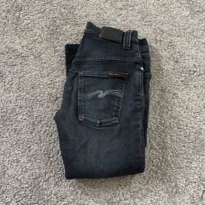 Mörkblå Nudie Jeans 27/32 - Snygga mörkblå jeans från Nudie Jeans med klassisk femficksdesign och cool broderad detalj på bakfickan. Modellen har normal passform och är tillverkad i stretchig bomull för extra komfort. Perfekt för dig som gillar stilrena och tidlösa jeans.