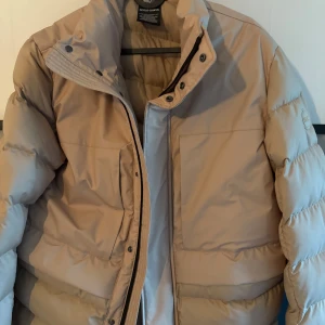 Beige pufferjacka från Colmar - Snygg beige pufferjacka från Colmar med hög krage och diskret logga på ärmen. Jackan har dragkedja och tryckknappar framtill, samt två stora fickor. Tillverkad i polyester från Recycled Essential-kollektionen. Perfekt för kalla dagar och riktigt trendig stil. Ny pris ca 6 tusen