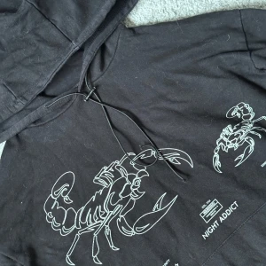 Svart hoodie Night Addict - Svart hoodie från Night Addict med stor vit skorpionprint på framsidan och coola grafiska detaljer. Storlek XS. Tröjan har huva med snörning, lång ärm och en klassisk magficka. Perfekt för dig som gillar streetwear och vill sticka ut med en unik design.