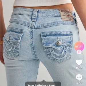True Religion ljusblå bootcut jeans - Säljer ett par ljusblå jeans från True Religion med ikoniska broderade fickor och silverfärgade knappar. Modellen har låg midja och bootcut-ben, vilket ger en snygg siluett. Jeansen är tillverkade i klassiskt denim och har detaljerade sömmar på bakfickorna.