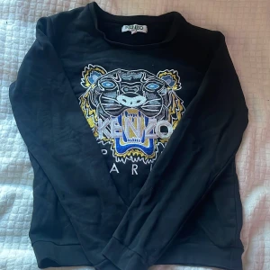 Svart Kenzo sweatshirt med tigerbroderi - Svart sweatshirt från Kenzo med ikoniskt tigerbroderi i blått, gult, vitt och grått på bröstet. Tröjan har rund hals och långa ärmar, och är tillverkad i mjuk bomull.