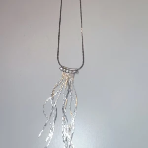 Silvrigt halsband med glittriga detaljer - Unikt halsband i silverfärg med glittriga stenar på en böjd stav och flera tunna, vågiga kedjor som hänger ner. Perfekt för dig som gillar statement-smycken med lite extra bling och rörelse.