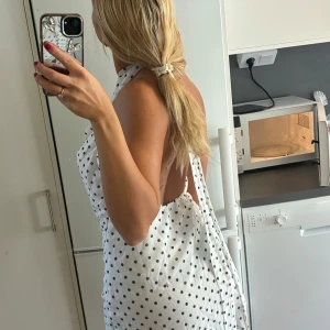 Vit prickig kortklänning med öppen rygg - Säljer en vit kortklänning med svarta prickar och halterneck. Klänningen har en snygg öppen rygg och ett draperat fall framtill. Perfekt för sommarens varma dagar, lätt och luftig i materialet. Passar dig som gillar en trendig och lekfull stil.