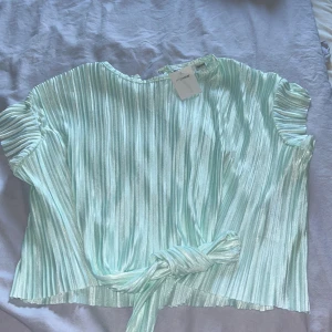 Plisserad mintgrön topp från MyWear - Snygg plisserad topp i mintgrönt från MyWear med korta ärmar och knytdetalj framtill. Materialet har en skimrande finish som ger en cool vibe och toppen har en rund halsringning. Perfekt för dig som gillar att sticka ut med färg och struktur.
