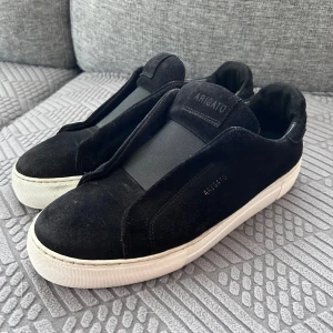 Arigato laceless  - Snygga svarta sneakers från Arigato med stilren design och vit platt sula. Skorna är tillverkade i mjuk mocka och har elastiska paneler istället för klassisk snörning, vilket ger en clean och modern look. Perfekta för dig som gillar minimalistisk stil.