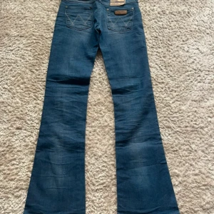 Wrangler Megan Bootcut jeans blå - Snygga blå jeans från Wrangler, modell Megan Bootcut med super låg midja. Klassisk femficksdesign, ljusblå tvätt och tydliga sömmar på bakfickorna. Materialet är jeans med en mjuk känsla och passformen är bootcut för en cool siluett. Midjemått - 33cm innerbenslängd - 79cm