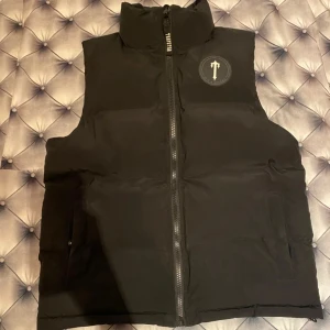 Trapstar - Trapstar dunväst i svart med hög krage och grov dragkedja framtill. Snyggt broderat Trapstar-tryck på ryggen och rund logga på bröstet. Västen har två fickor med lock och är perfekt för lager-på-lager. Stilsäker och urban look. Pris kan diskuteras 