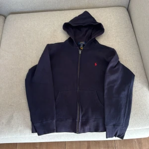 Mörkblå hoodie från Polo Ralph Lauren - Snygg mörkblå hoodie från Polo Ralph Lauren med klassisk röd broderad logga på bröstet. Tröjan har huva, dragkedja framtill och två praktiska fickor. Tillverkad i mjuk bomullsmix. Den är lite blekt. Passar dig som är runt 1,65 