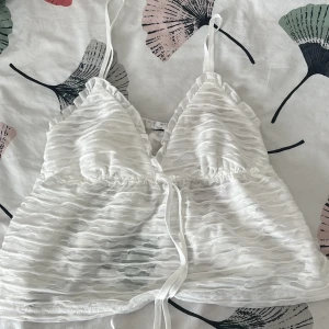 Vit omlott linnetopp med volang - Supersöt vit linnetopp med volangkant och justerbara smala axelband. Toppen har omlott-design och knytband framtill, samt ett vågigt, transparent mönster i tyget som ger en cool struktur. Perfekt för sommarens alla häng och chill. Priset kan även diskuteras❣️