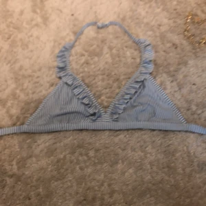 Blåvit randig bikiniöverdel med volang - Supersöt bikiniöverdel i blått och vitt med smala ränder och volangdetaljer längs kanten. Triangelformad modell med knyt i nacken och praktiskt spänne i ryggen. Perfekt för strandhäng och poolparty i sommar! Materialet känns mjukt och stretchigt.