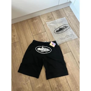 Svarta shorts med tryck från Yacht Club - Svarta shorts med elastisk midja och vit snörning. Framtill finns ett stort vitt tryck med motiv av en yacht i en oval ram. Materialet är mjukt och lätt polyester, perfekt för chill dagar. Snygg och enkel design som sticker ut med det marina temat. Vid snabb affär kan priset prutas