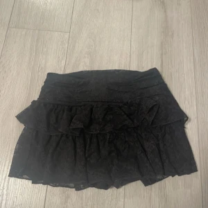Svart volangkjol med spets och dragkedja - Supersöt svart kortkjol med volanger och spetsdetaljer. Kjolen har en dold dragkedja i sidan och är fodrad med mjukt tyg under spetsen. Perfekt för dig som gillar en feminin och trendig look med extra volym.