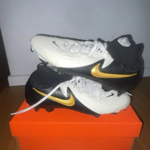 Nike fotbollsskor svart/vit/guld - Snygga Nike fotbollsskor med svart och vit ovandel, guldigt swoosh på sidan och strumpliknande skaft. Skorna har snörning och är tillverkade i syntetmaterial med textilinslag för extra komfort och passform. Perfekt för dig som vill sticka ut på planen. Använda 1 gång 