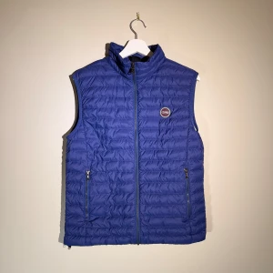 Blå dunväst från Colmar - Snygg blå dunväst från Colmar med quiltad design och dragkedja framtill. Västen har två praktiska fickor med dragkedja och en hög krage. Perfekt att slänga över en hoodie eller tröja för extra värme. Colmar-logga på bröstet ger en sportig vibe.