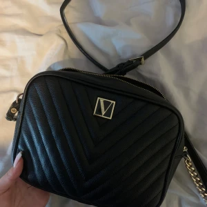 Svart axelväska från Victoria's Secret - Snygg svart axelväska från Victoria's Secret med quiltad design och guldfärgade detaljer. Väskan har en justerbar axelrem med kedja, dragkedja upptill och ett stilrent logomärke framtill. Perfekt storlek för det viktigaste och riktigt trendig look.