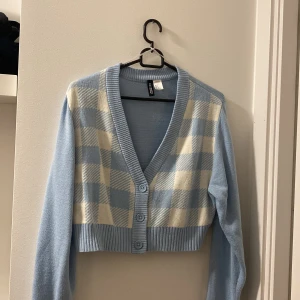 Blå rutig croppad kofta från H&M - Superfin croppad kofta från H&M Divided i ljusblått med rutigt mönster i blått och vitt framtill. Tröjan har långa ärmar, ribbad kant och tre knappar framtill. Perfekt för dig som gillar preppy och söt stil.