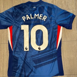 Chelsea Palmer #10 Nike fotbollströja S - Chelsea FC matchtröja med Palmer och nummer 10 på ryggen. Snyggt geometriskt mönster i olika blå toner, med vita och röda detaljer på sidorna. Officiell Nike Dri-Fit, klubbmärke på bröstet och Premier League-logga på siffrorna. Perfekt för dig som älskar fotboll och Chelsea.