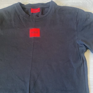 Svart t-shirt från Hugo Boss - Svart t-shirt från Hugo Boss med röd patch och HUGO-logga på bröstet. Klassisk rund hals och korta ärmar. T-shirten är gjord i mjuk bomull och har en clean, stilren look som passar perfekt till jeans eller shorts. Storlek S men passar även i folk med storlek M