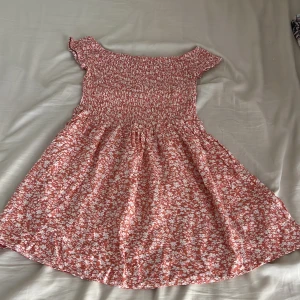 Rosa blommig kortklänning från SHEIN - Supersöt rosa kortklänning från SHEIN med vita blommor och smock upptill. Klänningen har korta ärmar och en flowy kjol som ger en chill vibe. Perfekt för varma dagar och har en offshoulder-look. Materialet är mjukt och lätt, troligen polyester.