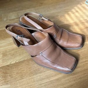 Vintage klackskor 39 - Säljer ett par bruna klackskor från JPW med fyrkantig tå och öppen häl. Storlek 39. Sparsamt använda. Små slitningar på baksidan.