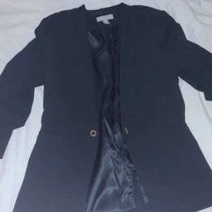 Svart kavaj från H&M, storlek 36 - Stilren svart kavaj från H&M i storlek 36. Klassisk rak passform med ett enkelt knappslut framtill och slät yta. Fodrad med glansigt svart tyg på insidan. Perfekt för dig som gillar minimalistisk och clean look.