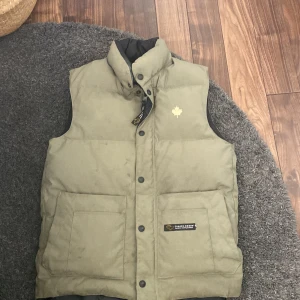 Olivgrön dunväst från Canada Goose - Säljer en olivgrön dunväst från Canada Goose med hög krage och stora fickor framtill. Västen har en diskret logga med lönnlöv på bröstet och svarta tryckknappar. Perfekt för kalla dagar och har en clean, klassisk look.