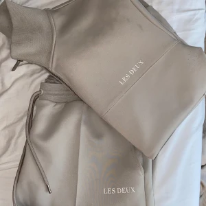 Beige set Les Deuw - Snygg beige set från Les Deux med half zip och byxor med diskret logga framtill. Perfekt för chill eller streetwear-stil. Finns dragsko i midjan för extra passform.