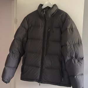 Svart pufferjacka från Polo Ralph Lauren - Snygg svart pufferjacka från Polo Ralph Lauren i storlek L. Jackan har hög krage, broderad logga på bröstet och flera dragkedjeförsedda fickor. Perfekt för riktigt vinterdagar  men också i höst 