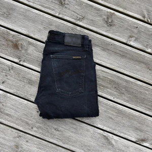 Svarta skinny jeans från Nudie Jeans - Säljer ett par svarta skinny jeans från Nudie Jeans med klassisk femficksdesign och snygga detaljer på bakfickorna. Jeansen har en mörk tvätt och är tillverkade i stretchig denim för skön passform. Perfekt för dig som gillar en smal siluett och stilren look. Kom gärna med frågor!🙌W30 L32