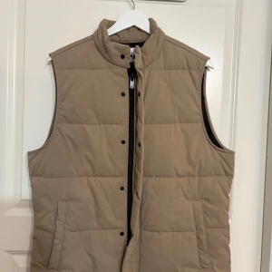 Beige quiltad dunväst från Zara - Snygg beige dunväst från Zara i storlek M. Västen har quiltad design, hög krage och både dragkedja och tryckknappar framtill. Två praktiska fickor på sidorna. Perfekt att slänga över en hoodie för extra värme och stil.