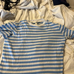 Blåvit randig croppad t-shirt H&M - Säljer en fräsch croppad t-shirt från H&M med blå och vita horisontella ränder. T-shirten har korta ärmar och rund halsringning. Perfekt för dig som gillar en chill och enkel stil. Materialet är mjuk bomull som känns skönt mot huden.