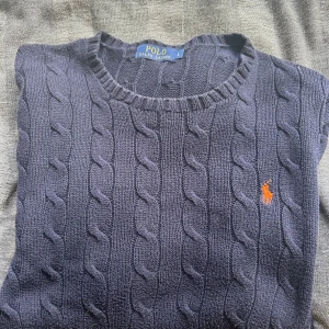 Mörkblå stickad tröja Polo Ralph Lauren - Snygg mörkblå stickad tröja från Polo Ralph Lauren med klassiskt kabelstickat mönster och orange broderad logga på bröstet. Rund halsringning och långa ärmar. Perfekt för dig som gillar preppy stil och vill ha något som känns både chill och stilrent. Är i storlek L men sitter mer som en M. Pris 199kr. Skick 7/10