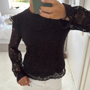 Svart spetsblus från Pieces XS - Svart långärmad blus från Pieces i storlek XS, helt i spets med snygga cirkelmönster och detaljerad kant vid hals och nederdel. Blusen har en liten knapp i nacken och transparenta ärmar med lätt puff. Perfekt för dig som gillar stilrena och unika plagg.