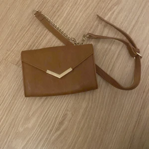 Brun kuvertväska från Beautiful You - Snygg brun kuvertväska med axelrem i skinnimitation och guldfärgad kedja. Väskan har ett stilrent V-format metallspänne framtill och flera fack inuti för kort och småsaker. Perfekt storlek för det viktigaste och enkel att matcha med olika outfits.