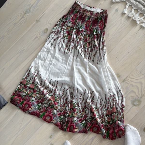 Vit långkjol med blommigt mönster VASA - Superfin långkjol från VASA TRUE i vitt med färgstarkt blommigt mönster i rött, grönt och rosa. Kjolen har smock i midjan för skön passform och är gjord i ett lätt och luftigt material, perfekt för varma dagar. Blommorna ger en romantisk och bohemisk vibe.