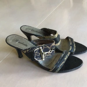 Svarta sandaler med guldmönster och klack - Snygga svarta sandaler från Leilida med guldmönstrade detaljer och metallspänne. Skorna har öppen tå, låg klack och en textur som liknar ormskinn. Perfekta för dig som gillar att sticka ut med coola accessoarer och detaljer.