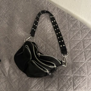 Axelremsväska - Cool svart axelväska med flera framfickor och silvriga dragkedjor. Axelbandet är dekorerat med vita prickar och har metallspännen. Perfekt för dig som vill ha en edgy och praktisk väska med plats för allt viktigt.