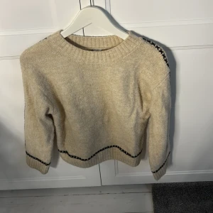 Beige stickad tröja med svarta detaljer - Säljer en beige stickad tröja i barnstorlek med svarta korsstygnsdetaljer längs ärmar och nederkant. Tröjan har rund hals och långa ärmar, perfekt för kyliga dagar. Enkel och stilren design som passar till det mesta.