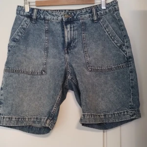 Blå jeansshorts med fickor från H&M - Blå jeansshorts från H&M med loose passform och flera stora fickor både fram och bak. Klassisk denimlook med ljus tvätt och robust känsla. Tillverkade i 100% bomull. Midwaist och med hällor för bälte. Stl 38