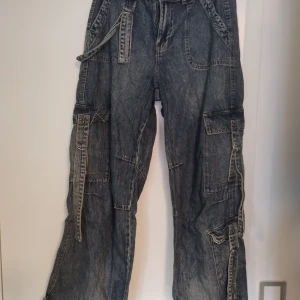 Baggy cargopants i mörk denim - Säljer ett par baggy cargopants i mörkblå denim med flera stora fickor och straps. Byxorna har snörning vid bensluten och en loose passform. De är lite slitna vid benslutet men endast använda ett fåtal gånger. 