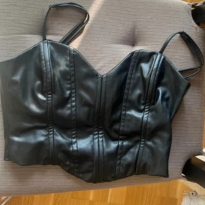 Svart korsett topp från Zara - Snygg svart korsett-topp med justerbara smala axelband och markerade sömmar framtill. Topen är croppad och har dragkedja i sidan för enkel på- och avtagning. Materialet är skinnimitation som ger en cool och edgy vibe.