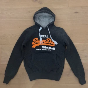 Mörkgrå Superdry hoodie med tryck - Snygg mörkgrå hoodie från Superdry med stort orange och vitt tryck på bröstet. Priset går att diskutera🙌