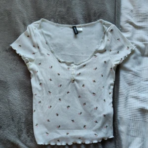 Vit blommig topp - En söt vit topp från h&m med små rosa blommor och v-ringad hals. Toppen har väldigt fina detaljer. En av mina gamla favoriter så den är använd men har inga tydliga defekter.