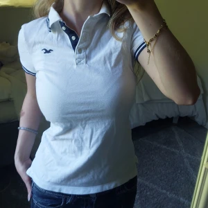 Vit piké från Hollister XS - Snygg vit piké från Hollister med marinblå detaljer på krage och ärmslut. Klassisk knappslå med tre knappar och broderad Hollister-logga på bröstet. Modellen är figurnära och har korta ärmar, perfekt till jeans eller shorts.