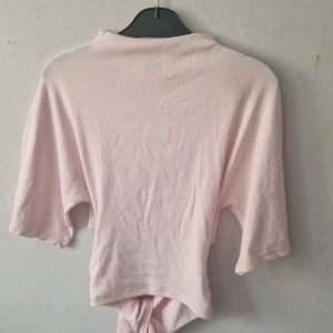 Ljusrosa croppad topp med knyt - Säljer en ljusrosa croppad topp med korta ärmar och snygg knytning baktill. Toppen har en enkel, stilren design och är gjord i mjuk bomull som känns skön mot huden. Perfekt för dig som gillar en chill och trendig look.